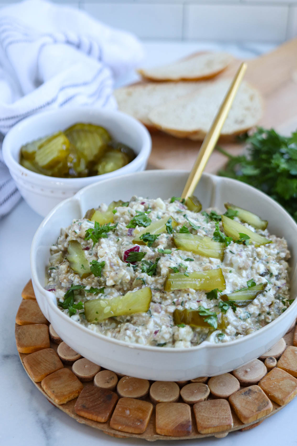 A bowl of creamy sardine salad dip garnished with sliced pickles and herbs sits on a coaster, with a spoon inside. Nearby are sliced pickles in a separate bowl and slices of bread on a wooden board.