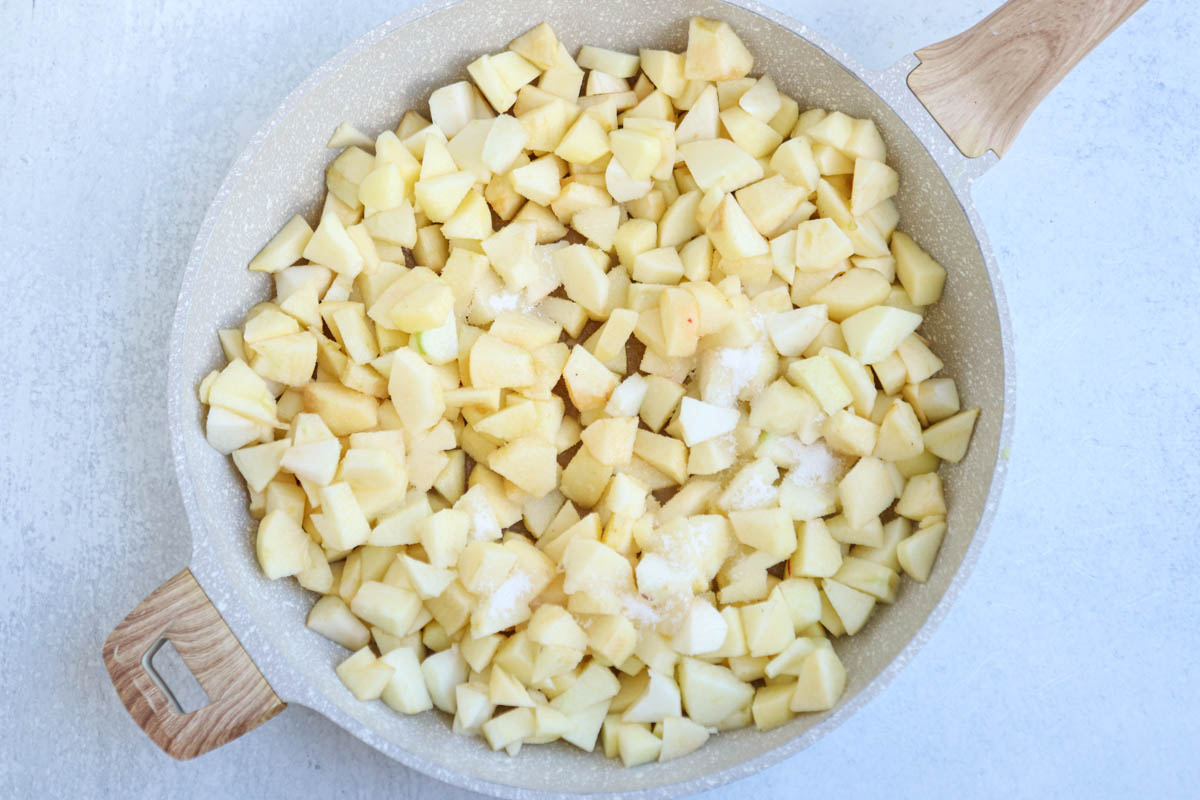 A beige skillet filled with chopped apples and a sprinkle of sugar, ready to be cooked for a classic Polish apple pie, sits on a light-colored surface.