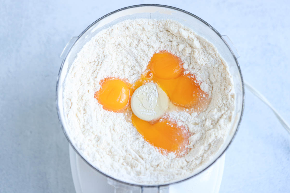 Top view of a food processor bowl filled with flour and four cracked eggs in the center, ready to be mixed on a light surface—perfect for preparing the dough for a classic Polish apple pie.