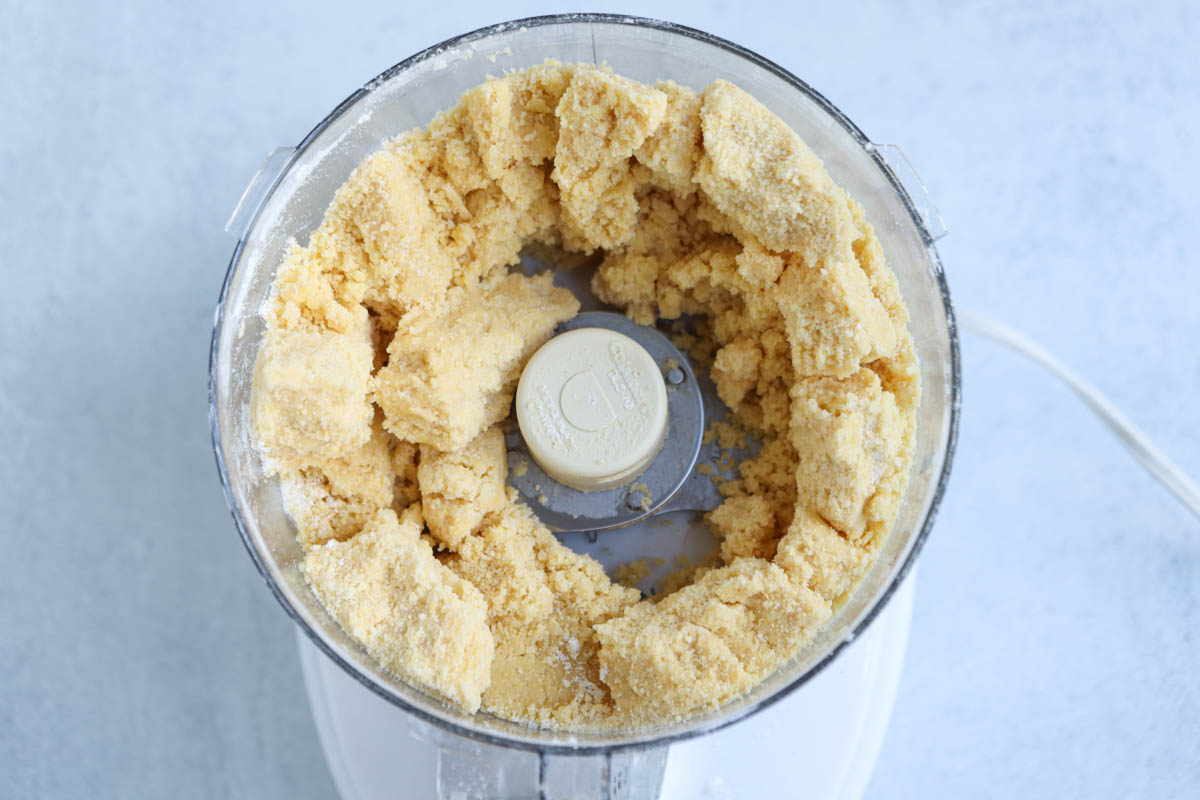 Top view of a food processor containing a crumbly, pale yellow dough mixture forming a ring around the blade—just the right texture for an authentic Polish apple pie—on a light-colored surface.