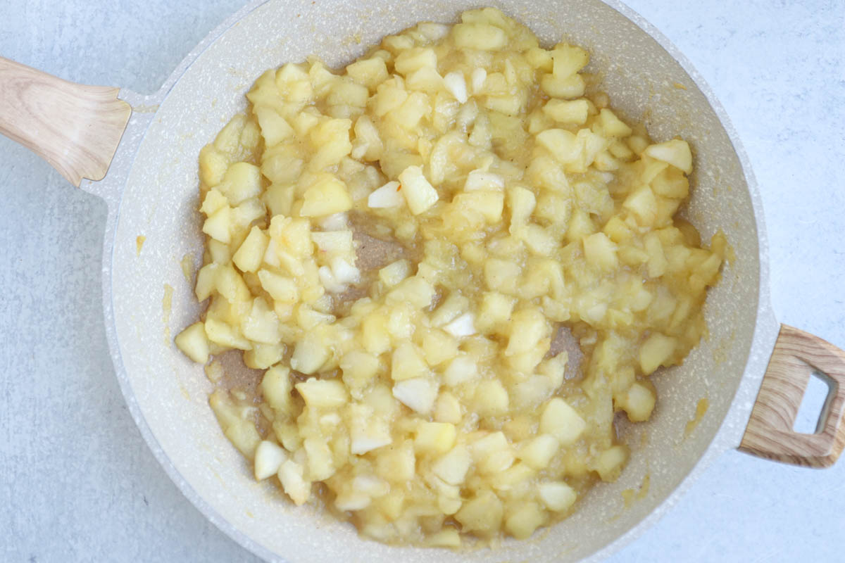 Chopped apples, soon to become the filling for a traditional Polish apple pie, are being cooked and softened in a light-colored frying pan with a wooden handle, set on a light gray surface.
