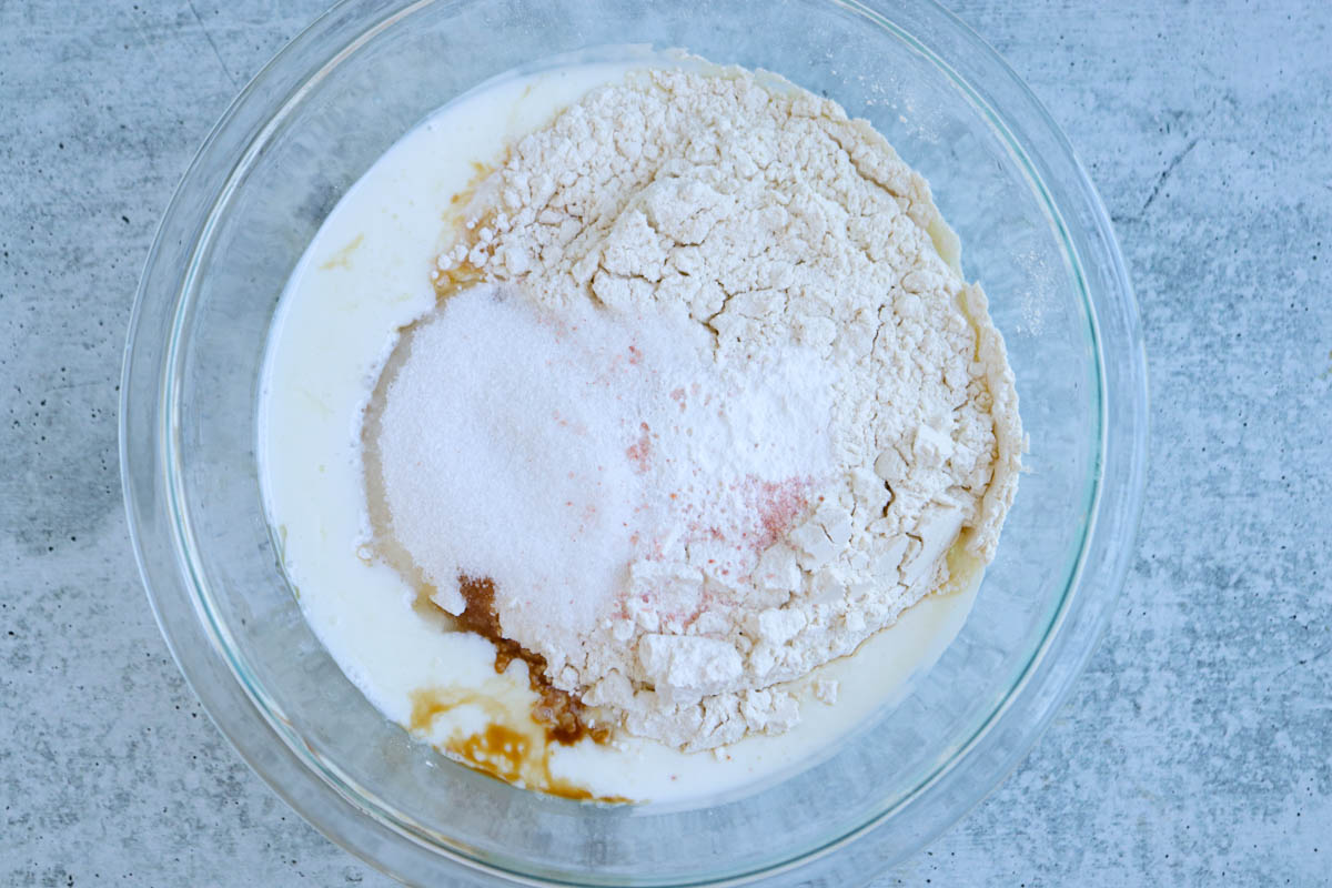 A glass bowl containing flour, sugar, baking powder, milk, and vanilla extract—ingredients ready to be mixed for traditional Polish apple pancakes—sits on a light gray countertop.