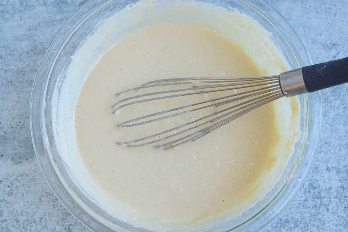 A metal whisk rests in a glass bowl filled with smooth, light beige batter for Polish apple pancakes on a gray countertop.