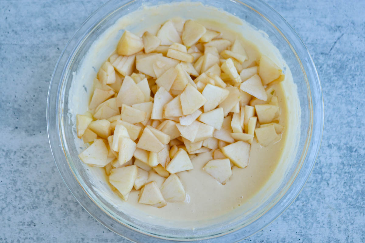 A glass bowl filled with cake batter for Polish apple pancakes and topped with chopped apple pieces, sitting on a light gray surface.