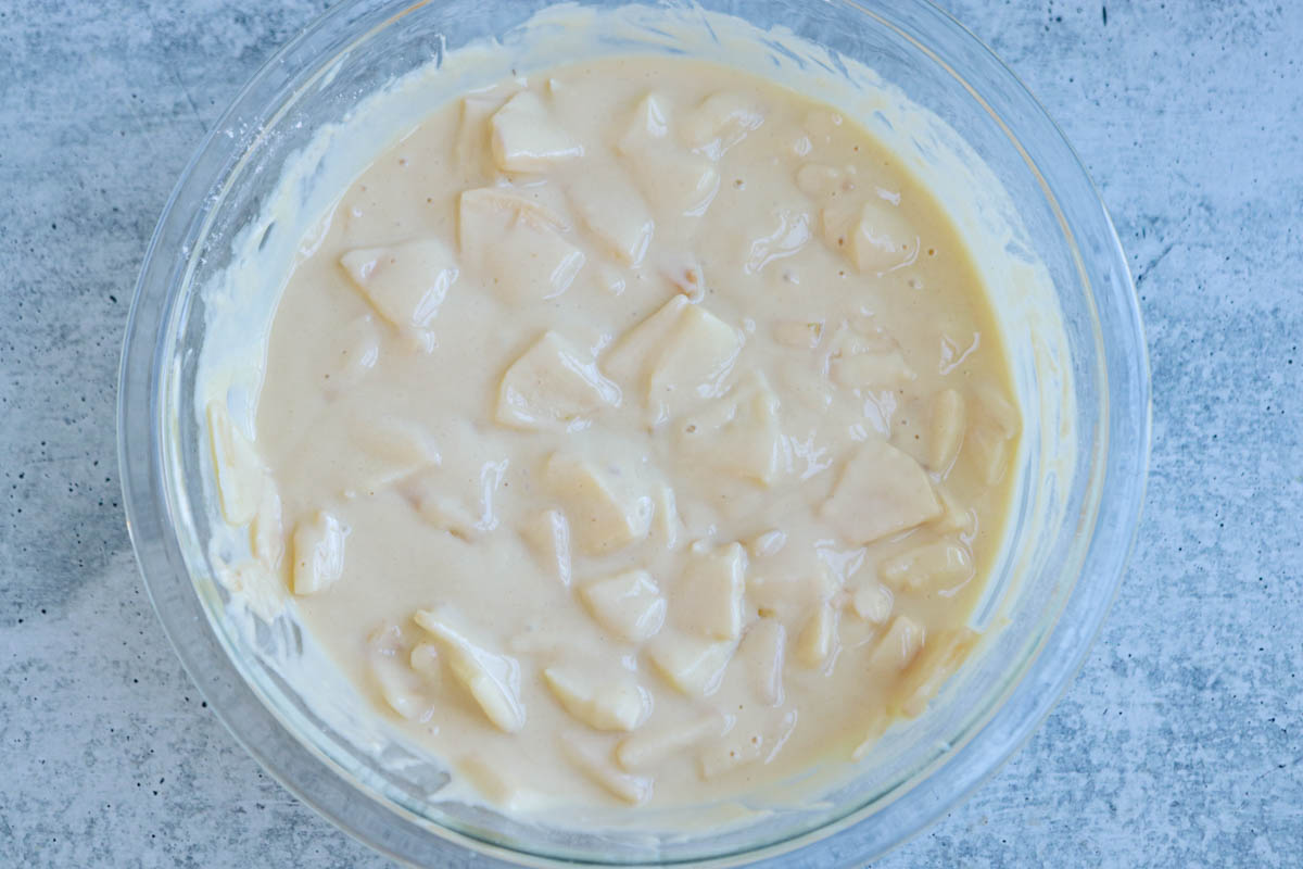 A clear glass bowl filled with creamy batter for Polish apple pancakes, mixed with chunks of sliced apples, set on a light gray, textured surface.