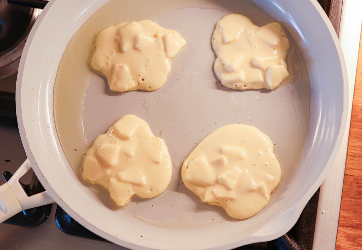 Four dollops of pancake batter with apple chunks—classic Polish apple pancakes—cooking in a white skillet on a stove, with oil visible around the pancakes.