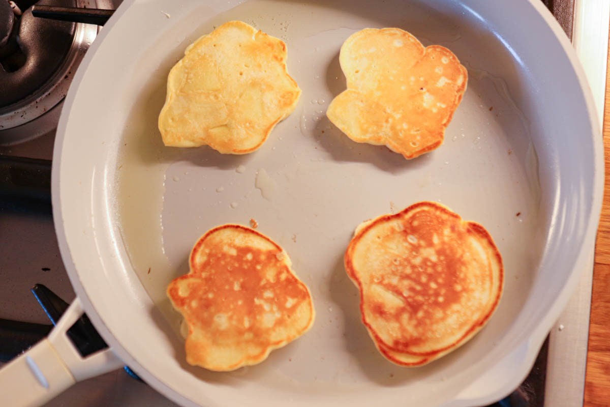 Four Polish apple pancakes cooking in a white nonstick skillet on a stovetop. The pancakes are golden brown and irregularly shaped, with tender apple slices inside, arranged evenly in the pan.