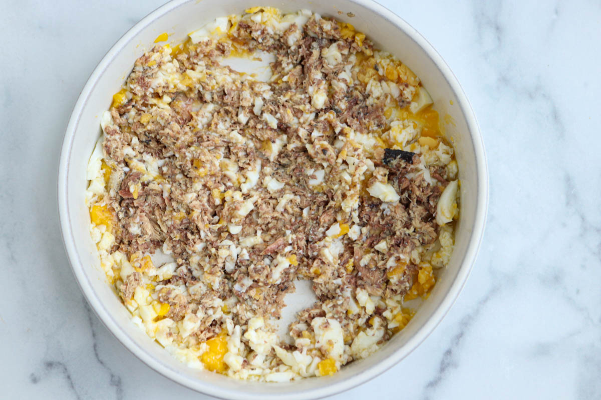 A white bowl on a marble surface contains a mixture of chopped hard-boiled eggs and flaked tuna, reminiscent of a classic sardine salad.