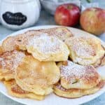 A plate of golden Polish apple pancakes, dusted with powdered sugar, with a jar labeled powdered sugar and two red apples in the background.