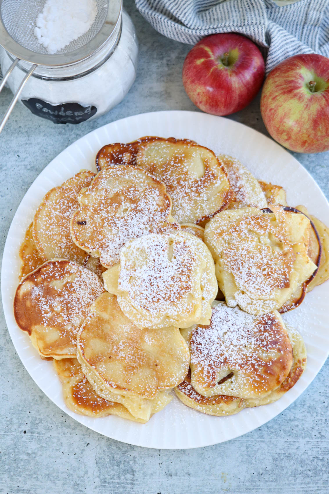A white plate filled with small, golden Polish apple pancakes dusted with powdered sugar. Two red apples and a jar of powdered sugar are nearby on a blue-gray surface.