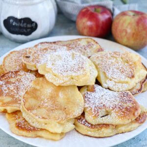 A plate of golden Polish apple pancakes, dusted with powdered sugar, with a jar labeled powdered sugar and two red apples in the background.