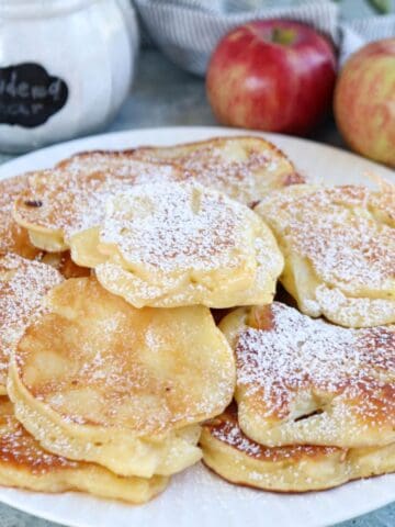 A plate of golden Polish apple pancakes, dusted with powdered sugar, with a jar labeled powdered sugar and two red apples in the background.