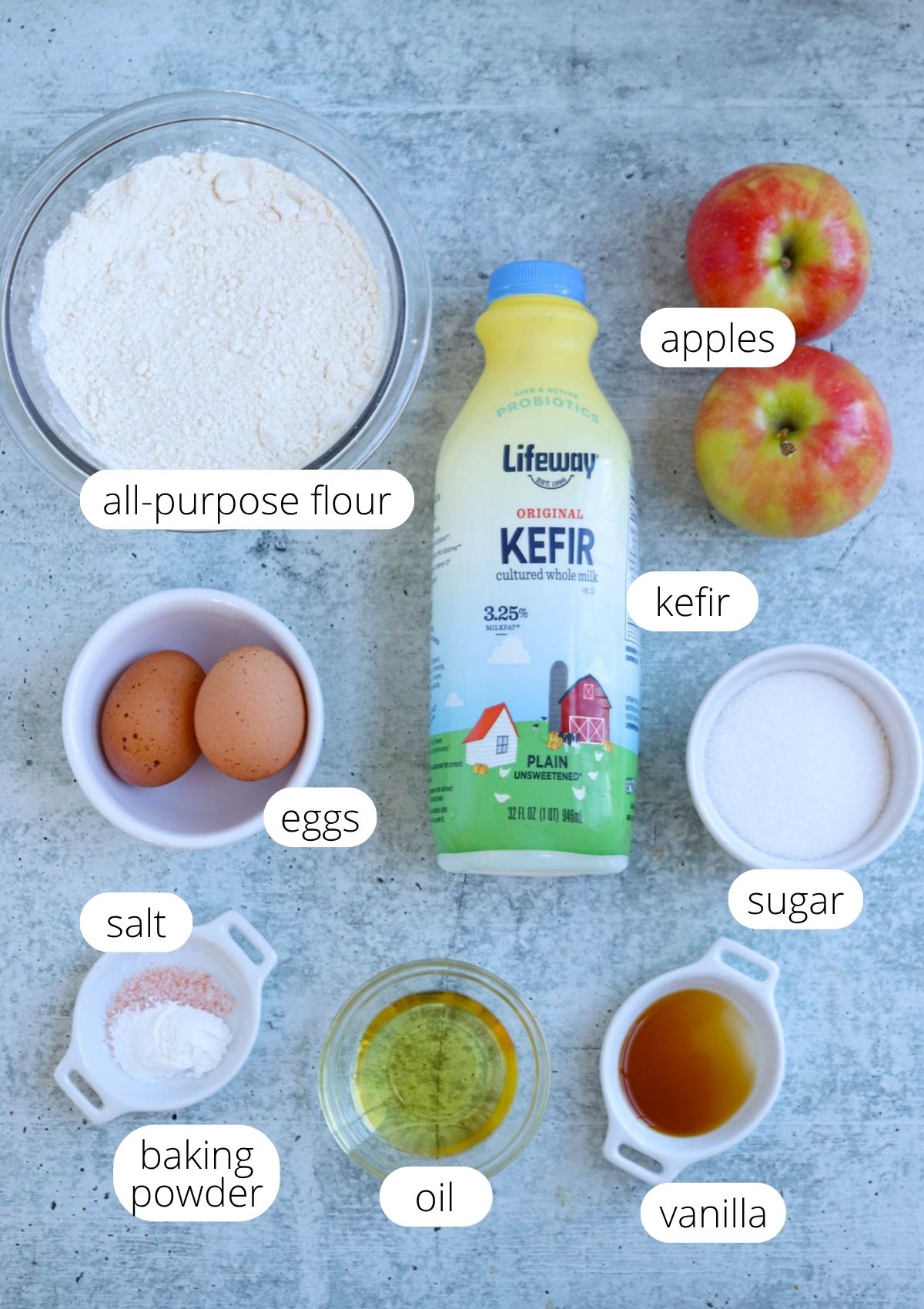 A flat lay of ingredients for baking Polish apple pancakes: apples, a bottle of kefir, all-purpose flour, eggs, sugar, salt, baking powder, oil, and vanilla—each in separate bowls and neatly labeled—arranged on a gray surface.