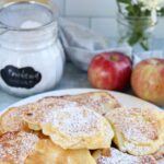 A plate of Polish apple pancakes dusted with powdered sugar sits on a countertop with apples, a jar of sugar, and flowers in the background. Text above reads: Polish Apple Pancakes. Racuchy z Jabłkami—a beloved traditional treat—are perfect for breakfast or dessert.