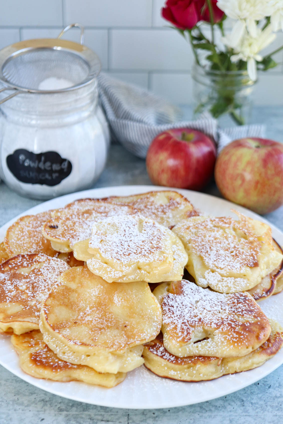 A plate of golden Polish apple pancakes dusted with powdered sugar sits on a table with red apples, a jar of powdered sugar, and a vase of flowers in the background.