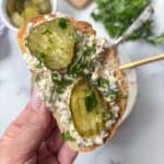 A hand holds a slice of bread topped with creamy sardine salad, two pickle slices, and chopped parsley. A bowl of pickles and fresh herbs are blurred in the background.