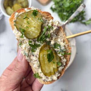 A hand holds a slice of bread topped with creamy sardine salad, two pickle slices, and chopped parsley. A bowl of pickles and fresh herbs are blurred in the background.