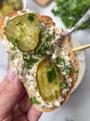 A hand holds a slice of bread topped with creamy sardine salad, two pickle slices, and chopped parsley. A bowl of pickles and fresh herbs are blurred in the background.