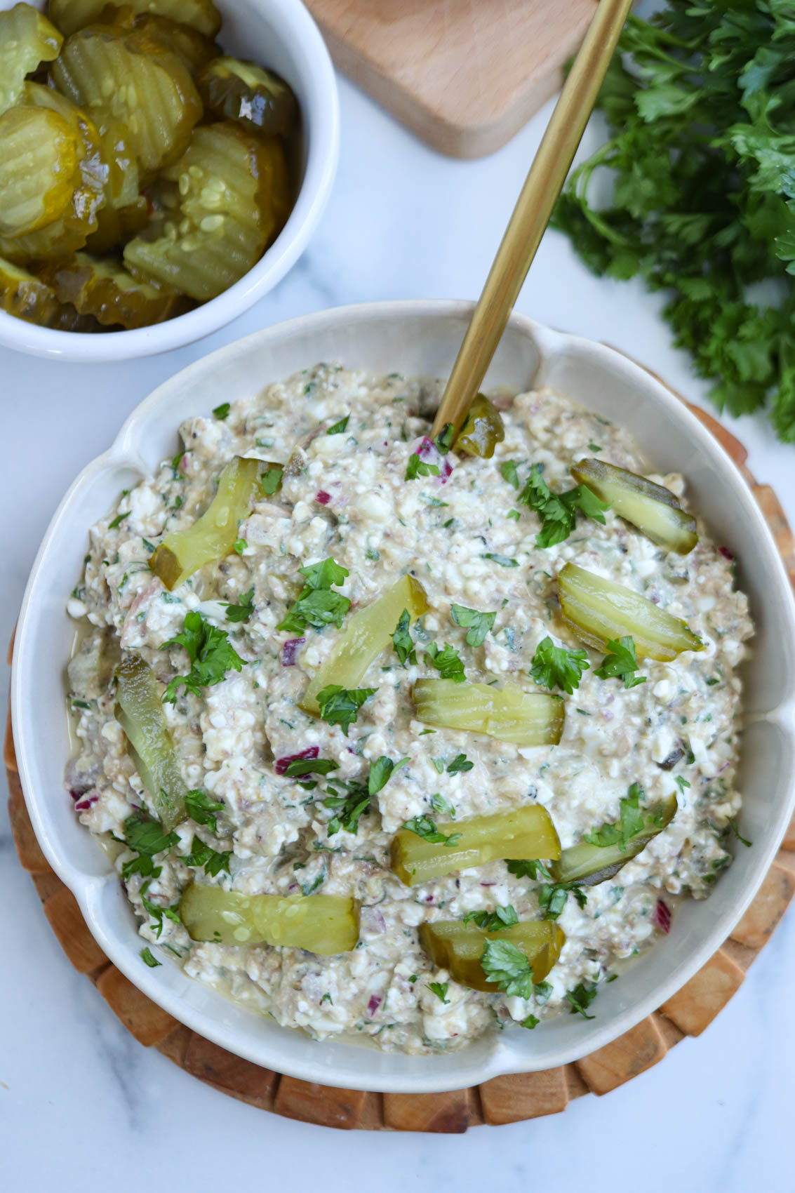 A bowl of creamy sardine salad topped with sliced pickles and chopped herbs sits on a wooden trivet. A fork rests in the bowl, and a small dish of pickles and fresh herbs are nearby.