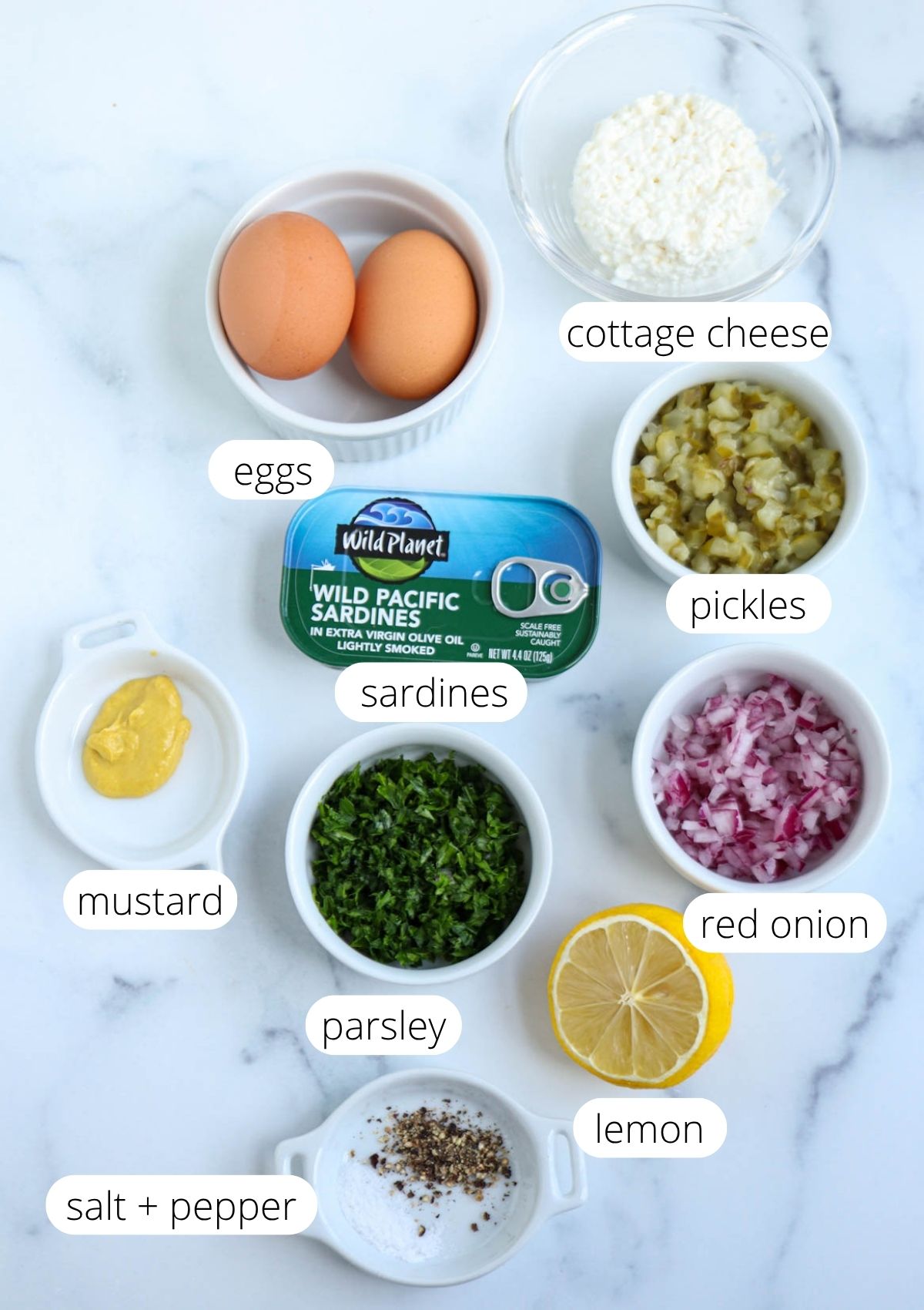 Ingredients for a sardine salad are arranged on a white surface: eggs, cottage cheese, pickles, canned sardines, mustard, parsley, red onion, lemon, and a small bowl of salt and pepper. Each ingredient is labeled to help you easily prepare this flavorful sardine salad at home.