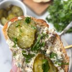 A hand holds a slice of bread topped with flavorful sardine salad, sliced pickles, and chopped parsley. In the background are fresh herbs, sliced bread, and a jar of pickles. Text: High Protein Sardine Salad—Super Nutritious.