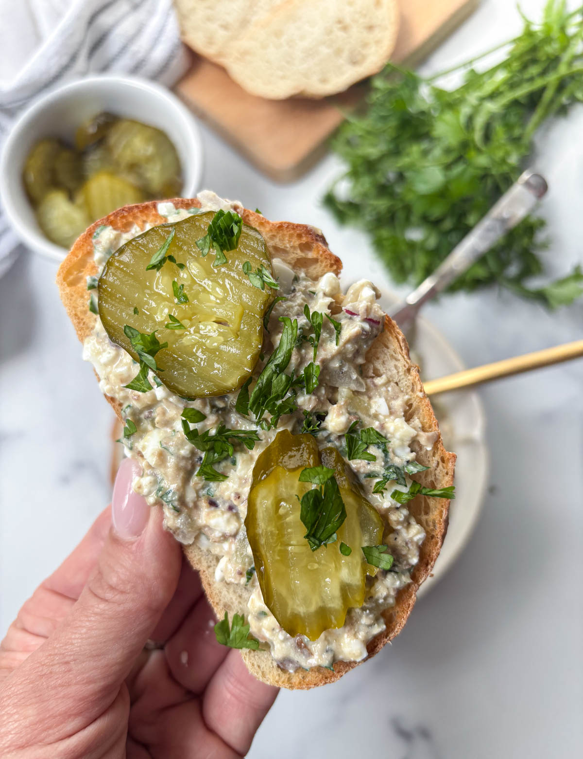 A hand holds a slice of bread topped with creamy spread, pickle slices, chopped parsley, and a generous spoonful of sardine salad. A bowl of pickles, fresh herbs, and a cutting board are blurred in the background.