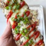 A hand holds a Polish zapiekanka, a toasted baguettetopped with melted cheese, mushrooms, ketchup, and sliced green onions.