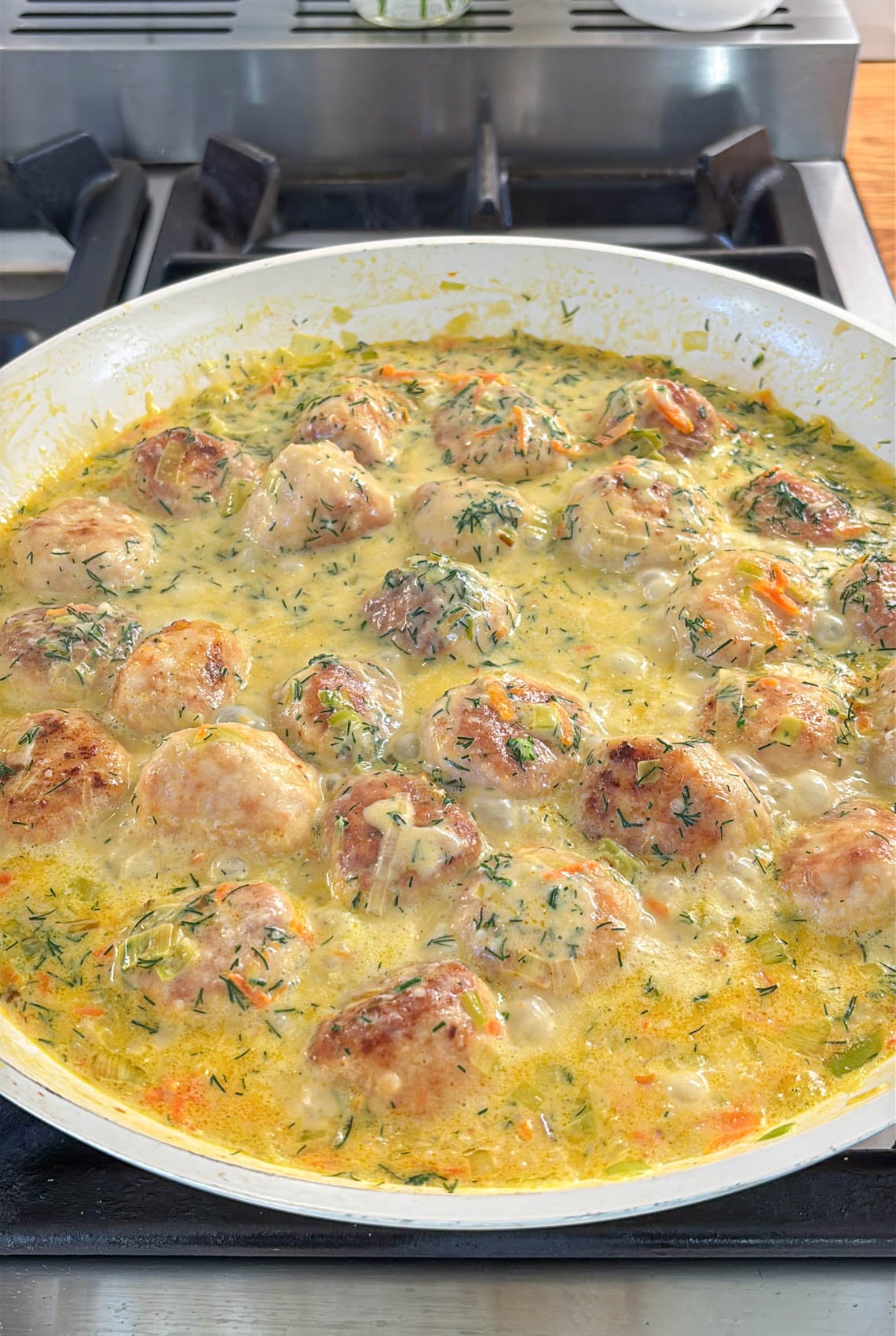Chicken meatballs simmering in a creamy, herb-flecked sauce with sautéed vegetables in a large pan on a stovetop.