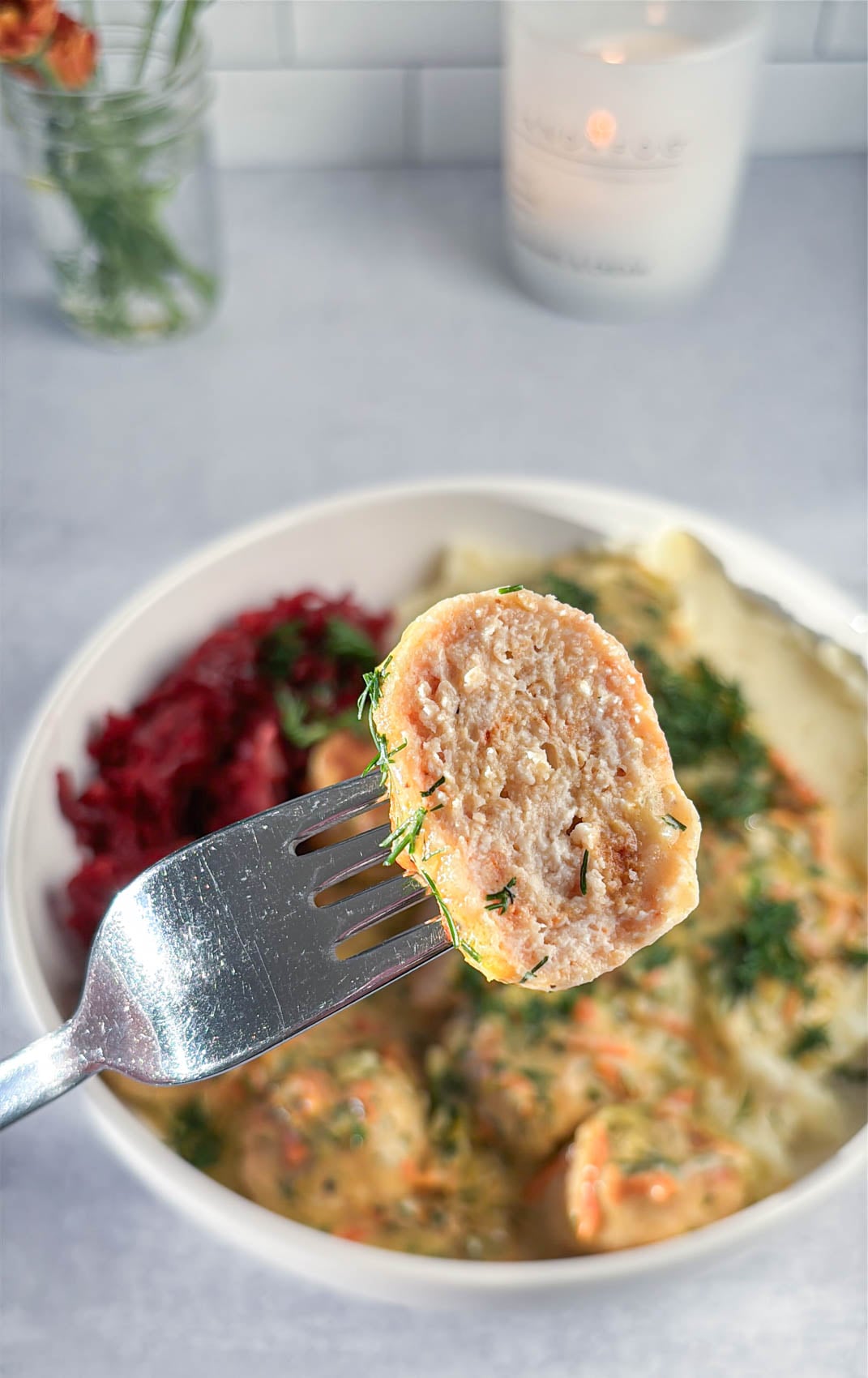 A fork holds a piece of a cooked, textured chicken meatball sprinkled with dill, with a bowl of similar meatballs and beet salad in the background.