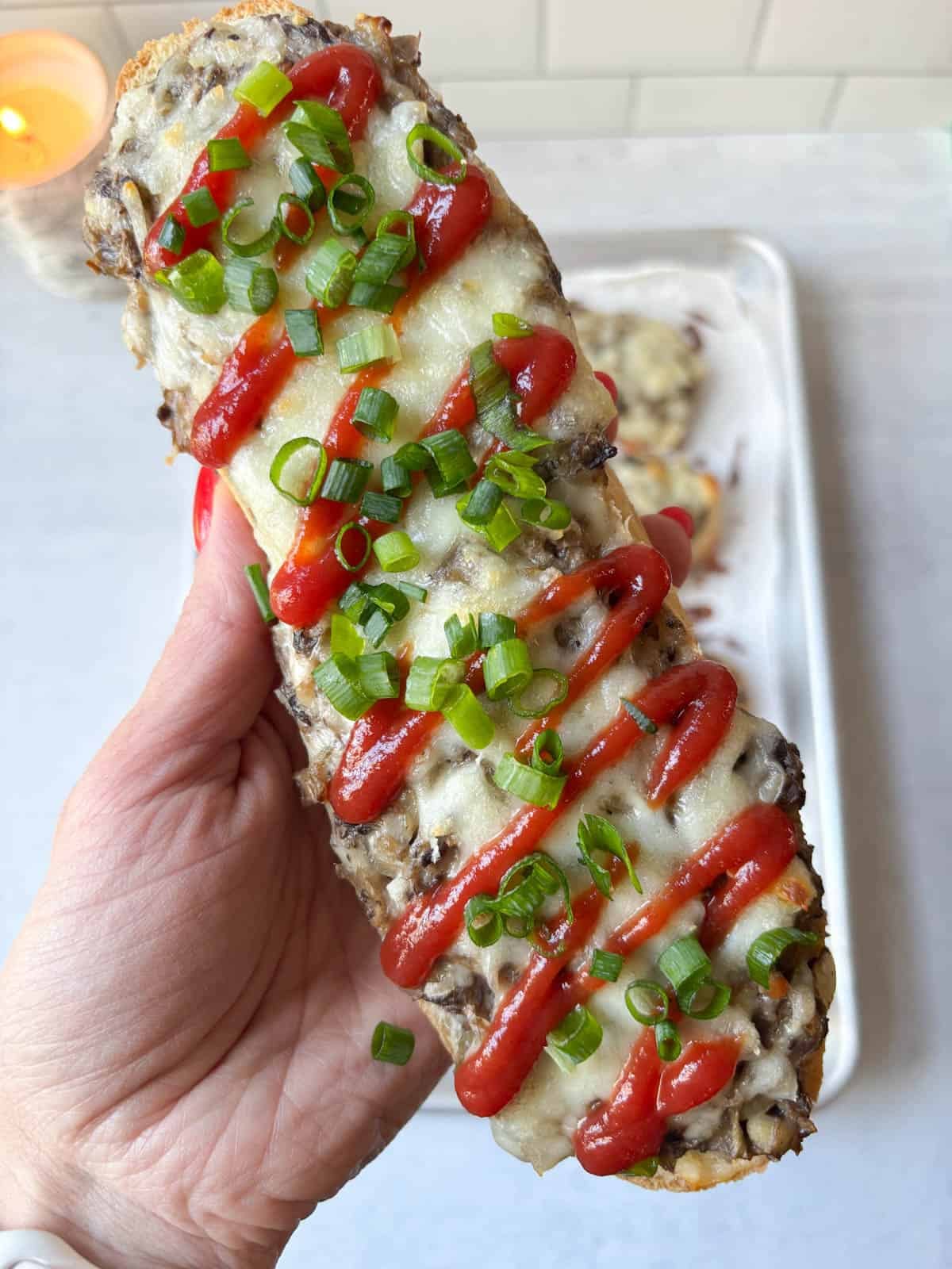 A hand holding a toasted baguette slice topped with melted cheese, savory mushrooms, a drizzle of ketchup, and chopped green onions.