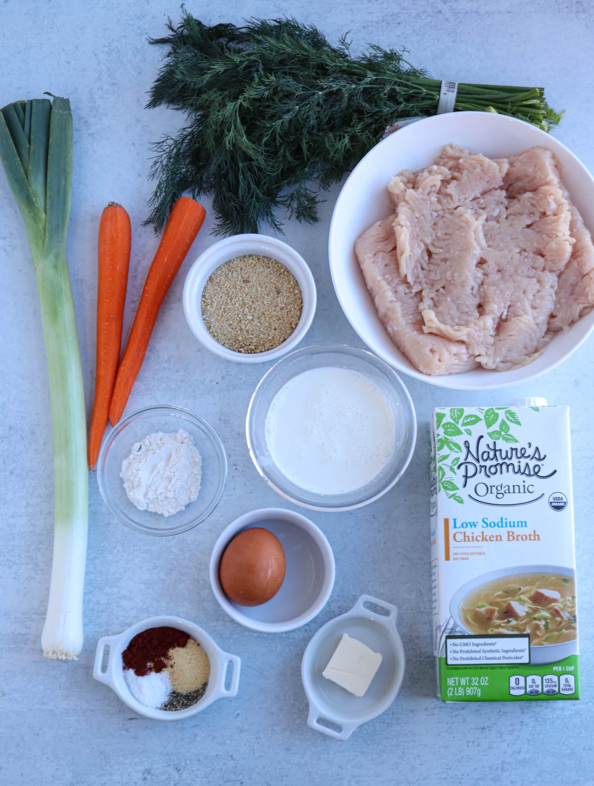 Ingredients for Polish chicken meatballs arranged on a counter: ground chicken, a leek, two carrots, fresh dill, breadcrumbs, flour, milk, an egg, spices, butter, and a carton of low-sodium organic chicken broth.