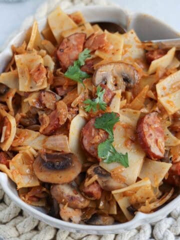 A bowl of pasta with sausage slices, mushrooms, and herbs, inspired by the classic Polish łazanki recipe, served on a textured mat with a fork placed in the bowl.