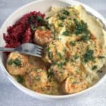 A bowl containing chicken meatballs in a creamy sauce, mashed potatoes, and shredded beets, garnished with fresh dill, with a fork resting in the dish.