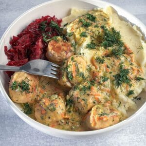 A bowl containing chicken meatballs in a creamy sauce, mashed potatoes, and shredded beets, garnished with fresh dill, with a fork resting in the dish.