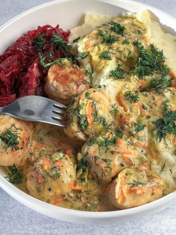 A bowl containing chicken meatballs in a creamy sauce, mashed potatoes, and shredded beets, garnished with fresh dill, with a fork resting in the dish.