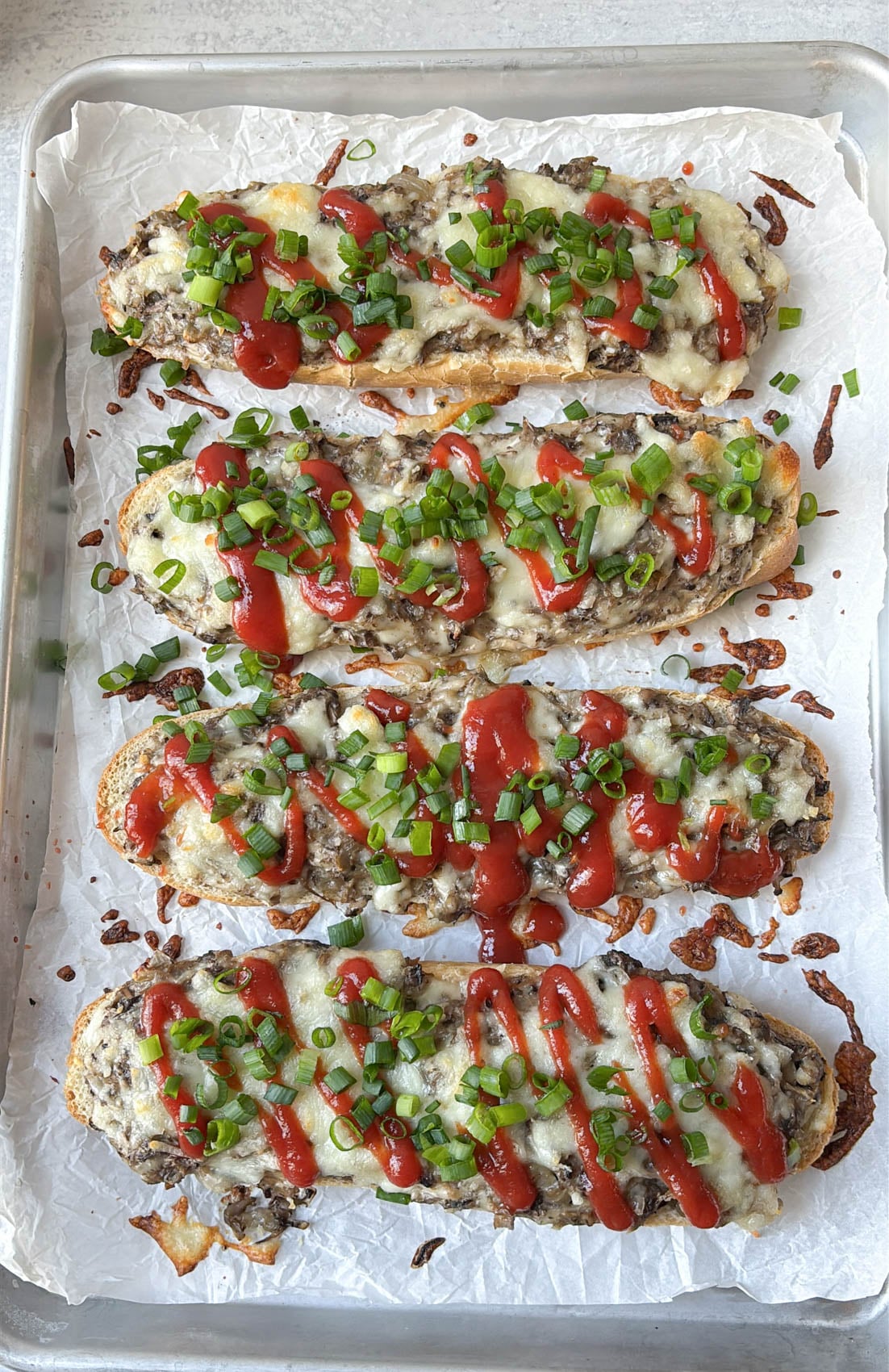 Four open-faced baguette sandwiches topped with melted cheese, a drizzle of ketchup, and chopped green onions on a parchment-lined baking sheet.