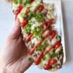 A hand holds an open-faced zapiekanka sandwich topped with melted cheese, drizzled ketchup, and sliced green onions.