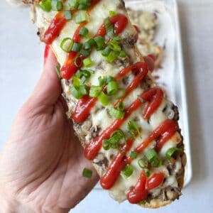 A hand holds an open-faced zapiekanka sandwich topped with melted cheese, drizzled ketchup, and sliced green onions.