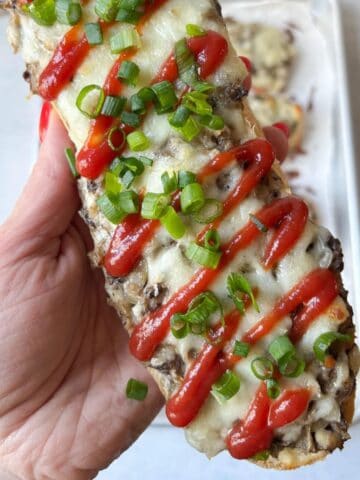 A hand holds an open-faced zapiekanka sandwich topped with melted cheese, drizzled ketchup, and sliced green onions.