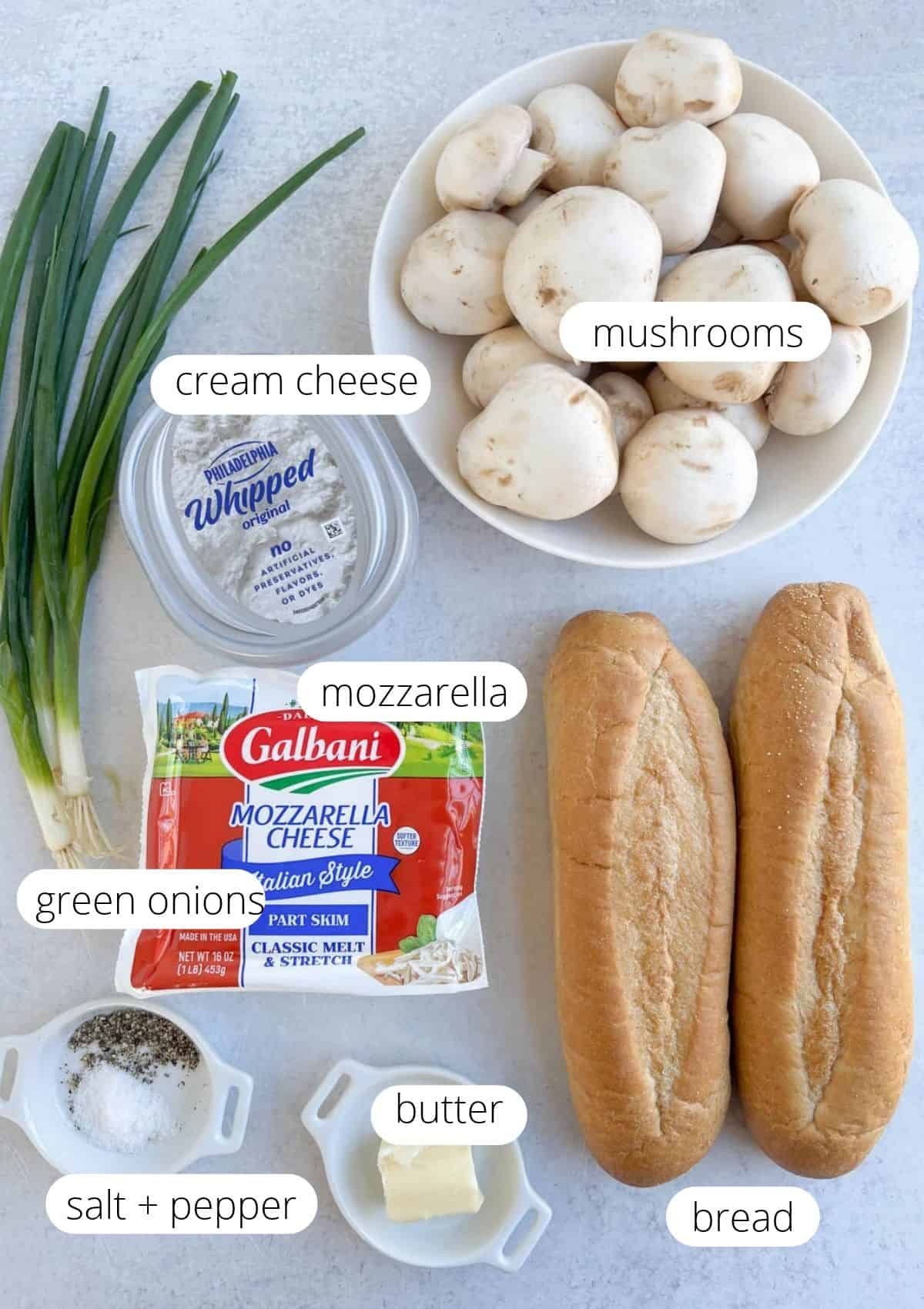 Ingredients for zapiekanka arranged on a counter: a bowl of mushrooms, green onions, a tub of whipped cream cheese, a package of mozzarella cheese, two loaves of bread, butter, salt, and pepper.